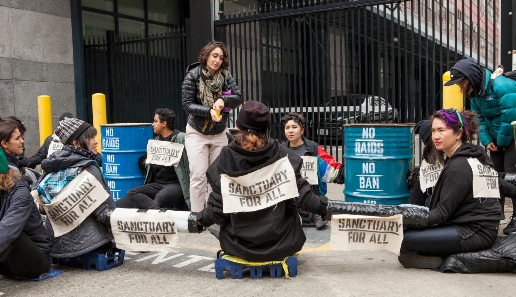 Protesters with locked arms and signs reading "Sanctuary for All" and "No Raids No Ban No Wall" block a driveway at a rally against Immigration and Customs Enforcement (ICE) in San Francisco, 28 Feb. 2018. Photo Pax Ahimsa Gethen, CC BY-SA 4.0.